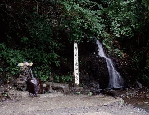 緑豊かな木々に囲まれ、岩肌を流れる滝の横に「丹沢名水 竜神の泉」の標柱が立ち、左に蛇口から甕へ水が注がれる水汲み場がある竜神の泉の写真