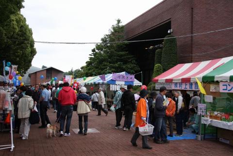 赤レンガの広場で開催されている秦野市商工まつりで、縞模様の屋台テントが立ち並び、犬を連れた人を含む多くの来場者が行き交っている写真