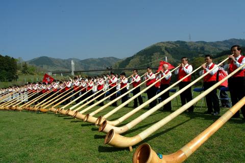 広々とした芝生の上で、赤いベストと白いシャツを着た大勢の演奏者たちが、アルプホルンと呼ばれる非常に長い木製の楽器を一斉に構えて演奏している写真