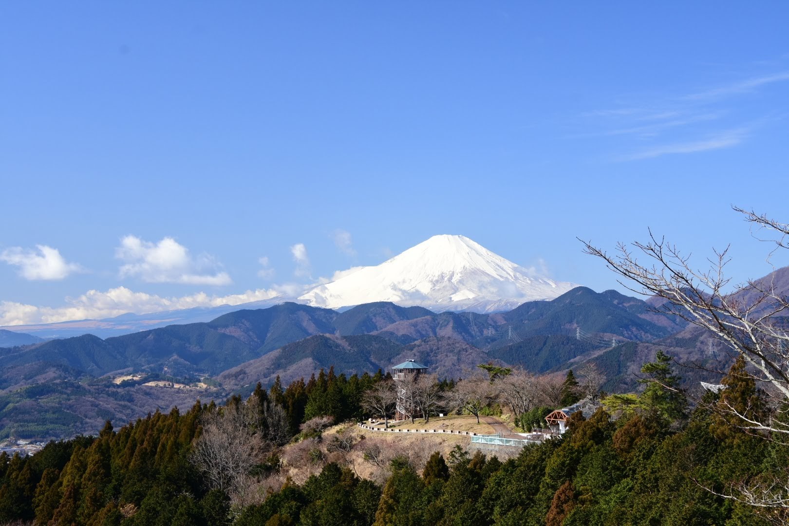 青空と白い雲を背景に、冠雪した富士山が中央にそびえ立ち、手前には茶色と緑の山並みと展望台のある公園施設、針葉樹と落葉樹が混在する冬の丹沢山地からの遠景写真