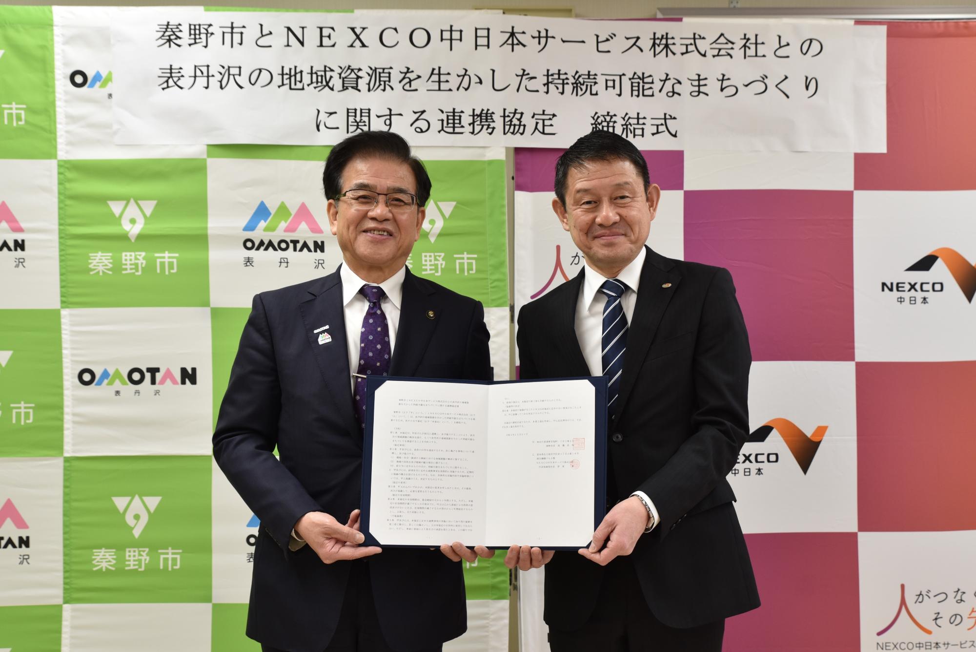 「秦野市とNEXCO中日本サービス株式会社との表丹沢の地域資源を生かした持続可能なまちづくりに関する連携協定 締結式」と書かれた背景幕の前で、高橋市長との男性が署名済みの協定書を開いて正面を向いて立っている