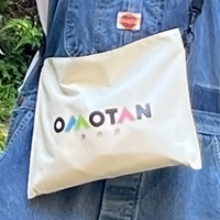 青いデニム生地の胸元に、白い布製バッグが斜めがけされており、そのバッグには虹色の文字で「OMOTAN」というロゴが印刷されているサコッシュの写真