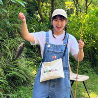 緑豊かな森林の前で、白い帽子と青いオーバーオール、白いTシャツを着た人物が笑顔で両手を上げてガッツポーズをしている写真