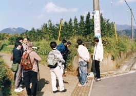 晴れた日に田舎道の角で、ハイキングやフィールドワークをしている数名のグループが、草木の生えた土手と電柱の前に集まり、地図か何かに目を向けながら説明を聞いている様子の写真