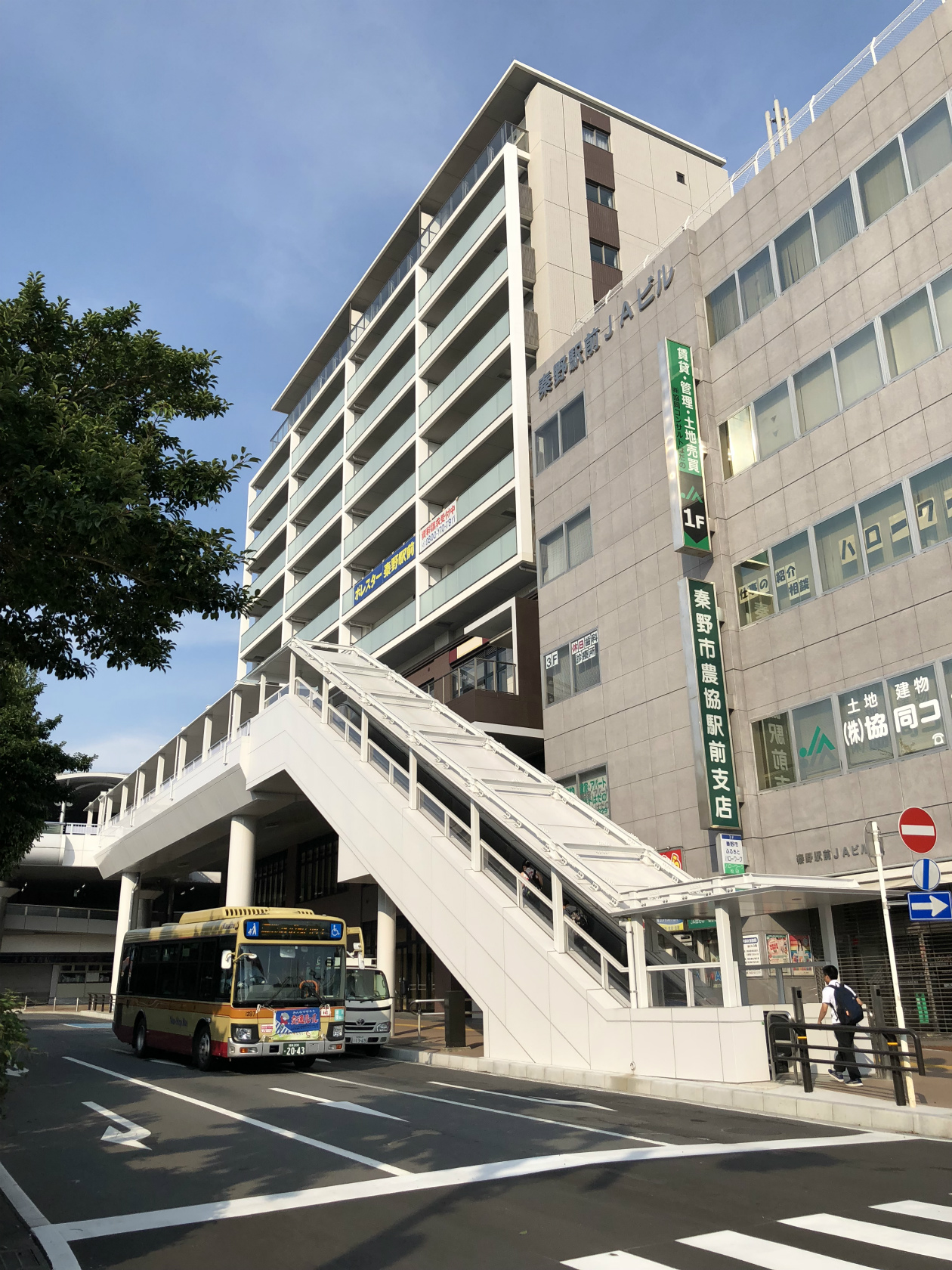 路線バスが横切る道路に面した歩道に白い屋根付きの歩道橋が設置され、その奥にオフィスビルと並んでマンションが建っている写真