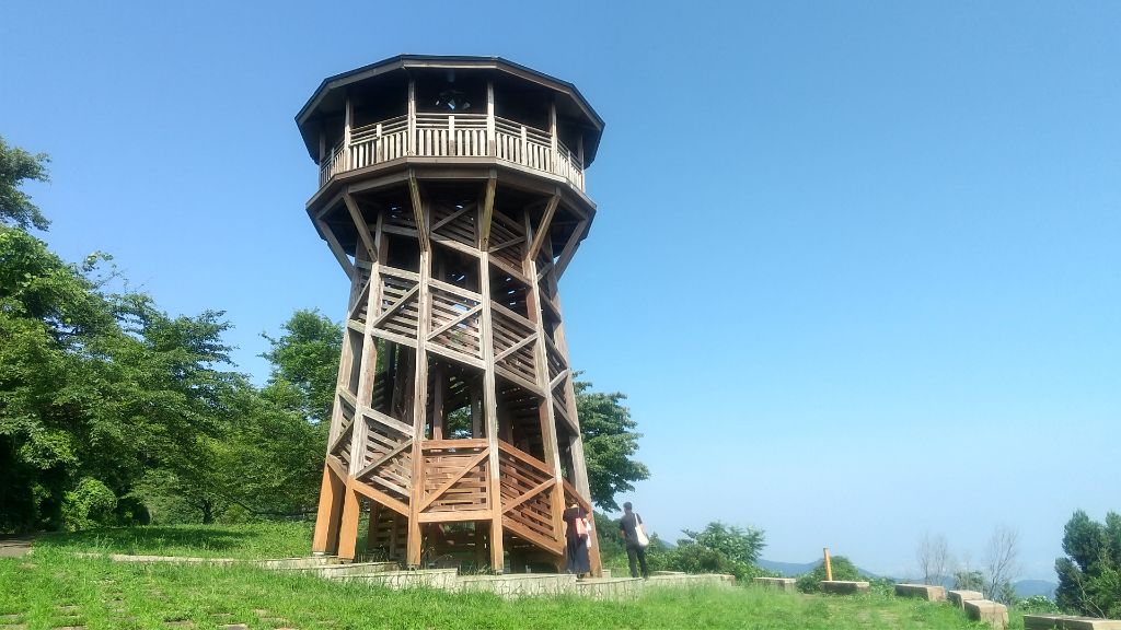 青空のもと、芝生の上に立つ木組みの複雑な構造を持つ八角形の展望台が下から見上げるように捉えられており、展望台の最上階には手すりが巡らされたスペースがあり、階段の脇には二人組の人物が立っている自然の中の風景写真