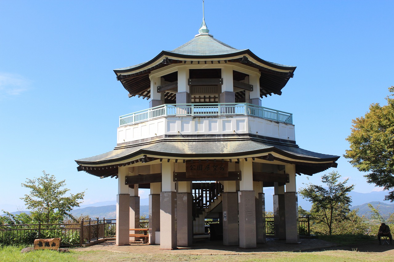 青い空を背景に、八角形の土台の上に建てられた二重の屋根を持つ展望台のような東屋が映されており、その建物の下部には「公園展望台」と書かれた看板があり、周囲には木々や山並みが広がる風景写真