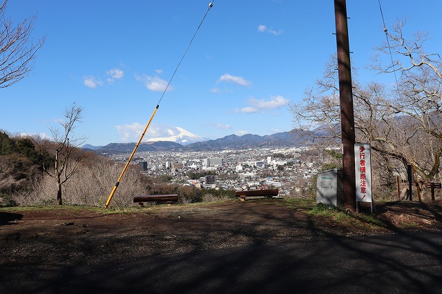 展望地から見た青空の下の風景で、雪を被った富士山が遠くの山並みの上にそびえ、眼下に広がる街並みが確認でき、手前の広場には木製のベンチが二つ設置され、「歩行者横断注意！」と書かれた赤い看板が見える広大な眺望の写真
