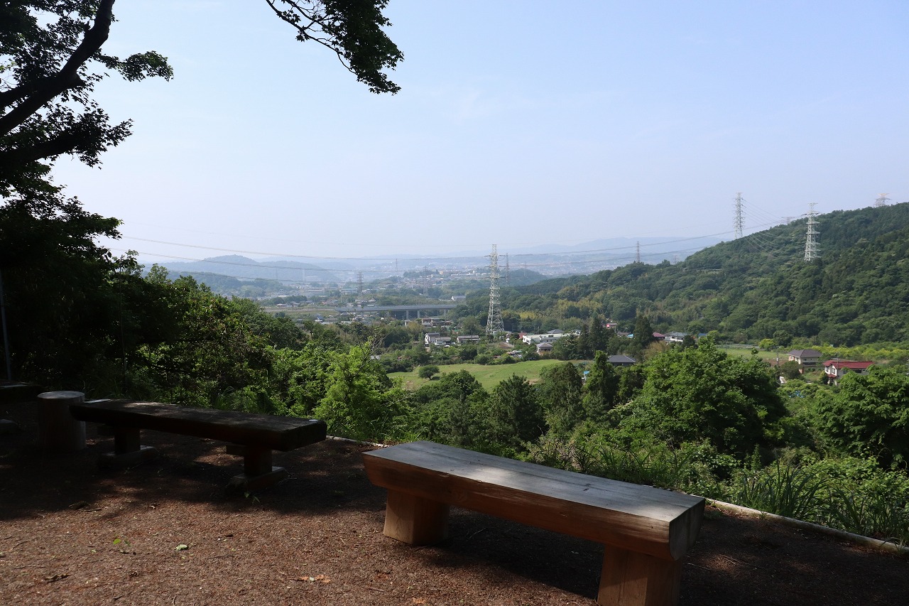 晴れた日の丘の上から、手前の土の広場に置かれた二つのシンプルな木製ベンチ越しに、青々とした森と谷間に広がる街並みを見下ろす風景が広がり、遠くの山肌には送電線用の大きな鉄塔が複数見える、自然の中の遠景写真