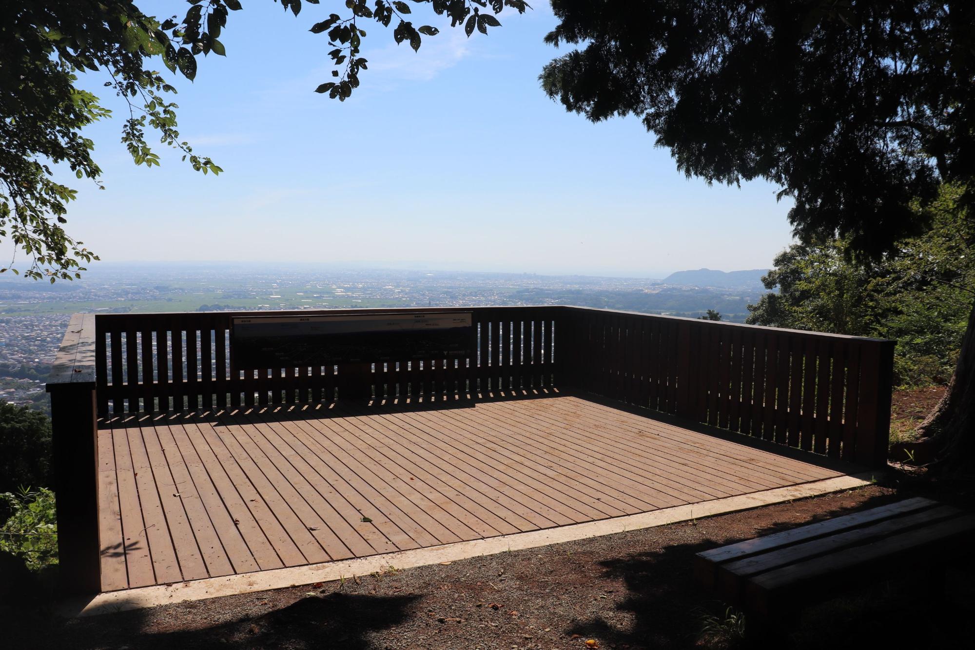 深い緑の木々に囲まれた展望台の全景で、木製の床が敷き詰められたデッキの周囲には、縦の板が並んだ木製の手すりが巡らされ、手すりの内側には展望案内板が設置されており、遠くには広大な市街地が広がる晴天の風景写真