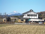白壁と黒い梁の大きな和風建物が建ち、その背後には雪をかぶった高い山が青空の下にくっきり見えている様子を田園風景に囲まれた建物を写した写真