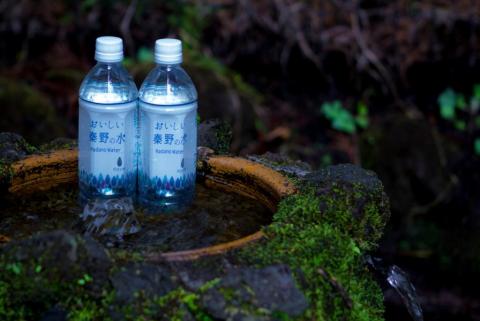 苔むした石の湧水口の前に、青いラベルに「秦野の水」と書かれた透明なペットボトル二本が並べられ、背景に緑の葉がぼやけて見える湧水の写真