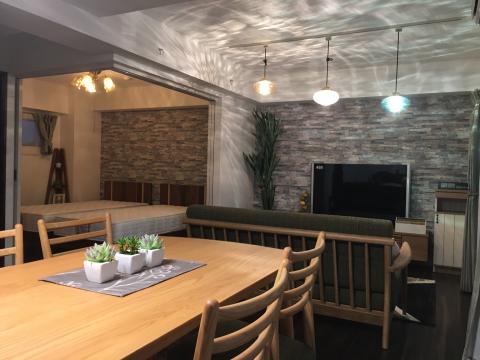 木製のダイニングテーブルと椅子が手前に配置され、背後に石目調のアクセントウォールと壁掛けテレビ、ペンダントライトが吊るされたモダンなリビングダイニング空間の写真