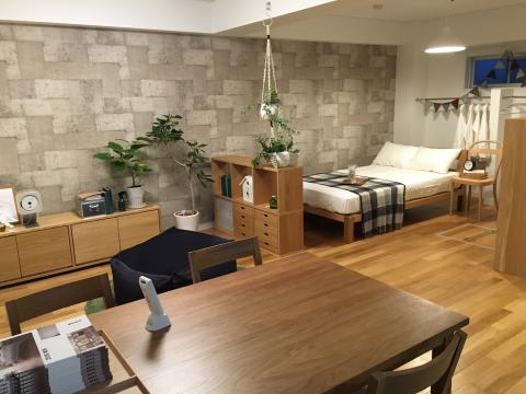 木製フローリングの床に、手前にダイニングテーブルと椅子、奥に木製ベッドフレームと収納家具、グレーのレンガ調アクセントウォール、複数の観葉植物が配置されたナチュラルテイストの室内の写真
