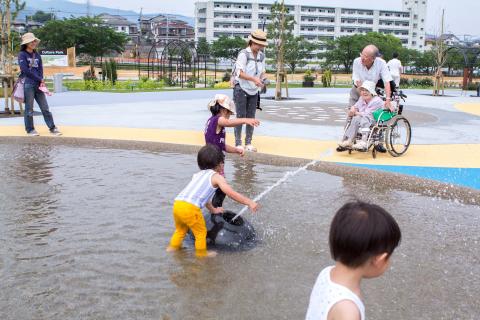 晴天の公園内にある浅い水遊び場で、黄色い長靴を履いた子どもたちや大人が水しぶきを上げて遊び、背後に遊具施設とマンション群が見える夏の親水広場の写真