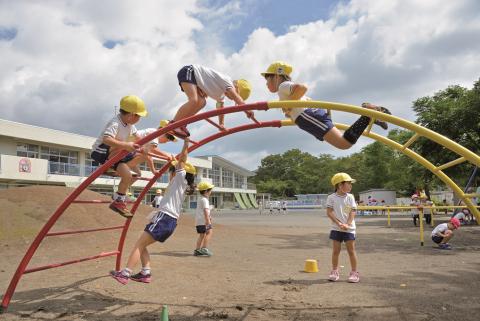 青空と白い雲の下、校庭に設置された赤と黄色のアーチ型ジャングルジムで黄色い帽子をかぶった複数の子どもたちが遊び、背後に学校の建物と緑の木々が見える写真