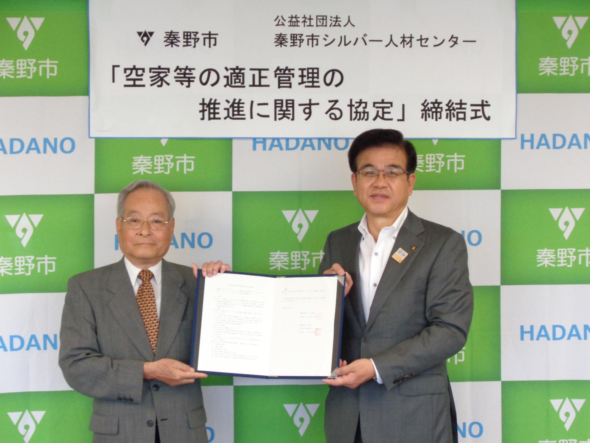 緑と白のロゴ入り背景幕の前で「空家等の適正管理の推進に関する協定締結式」と掲げられ、スーツ姿の秦野市長とシルバー人材センター代表が協定書を掲げる公式調印式の記念写真