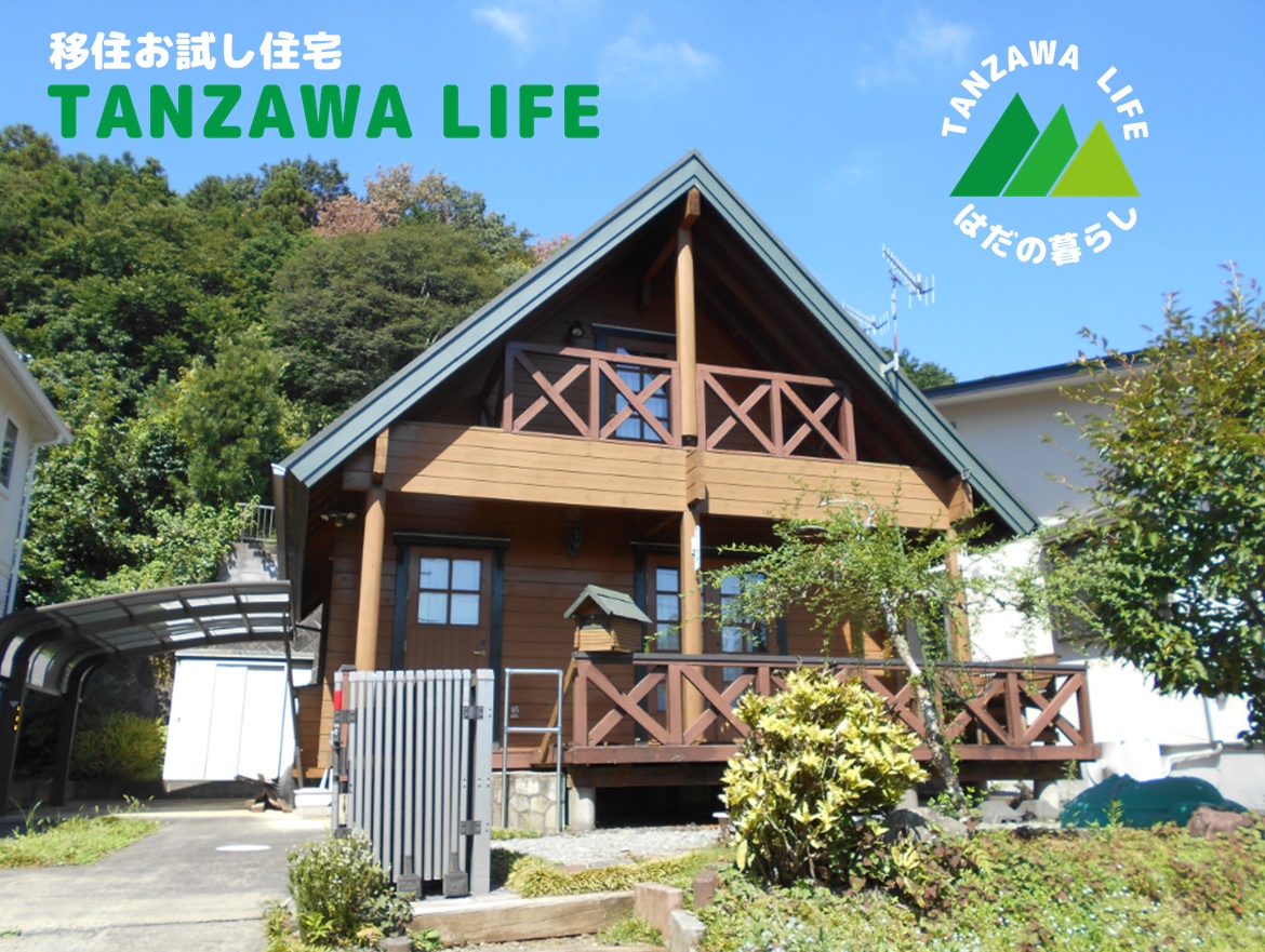 緑豊かな環境に建つ三角屋根の木造ログハウス風住宅の前に「移住お試し住宅 TANZAWA LIFE はだの暮らし」と緑の山ロゴを配した丹沢移住体験プログラムの紹介写真