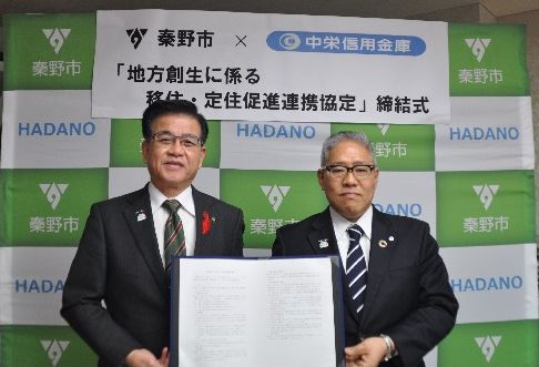 秦野市のロゴが並ぶグリーンのバックボードの前で、市長とスーツ姿の男性が協定書を掲げて記念撮影している締結式の写真