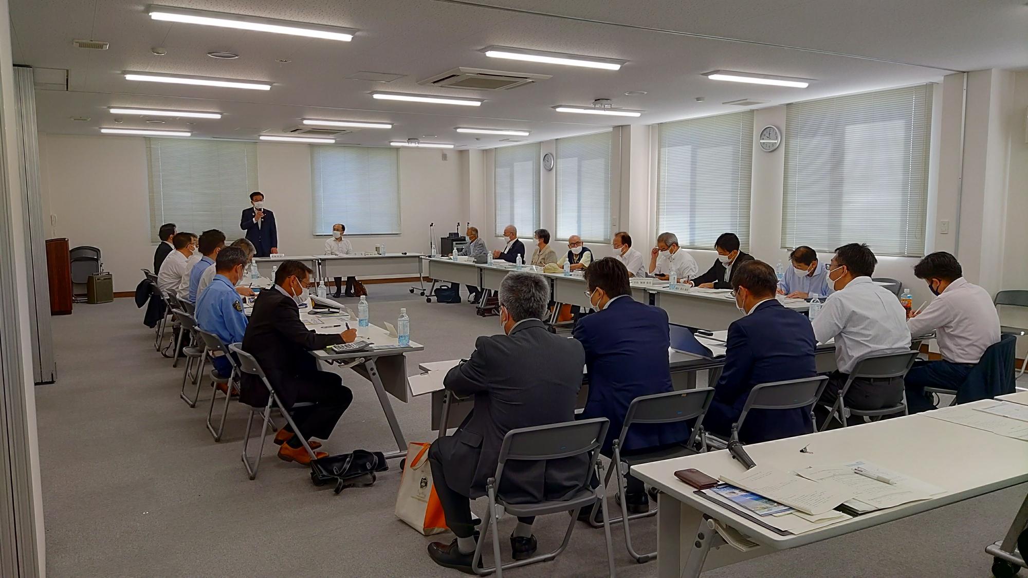 明るい蛍光灯と大きな窓のある白い会議室で、ロの字型に配置されたテーブルを囲む多数のスーツ姿の参加者が資料を手に会議が行われている写真