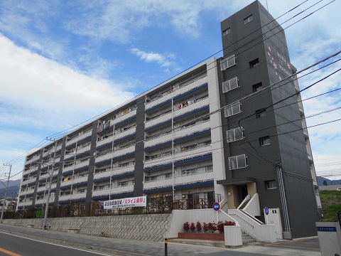 青空に浮かぶ白い雲を背景に、灰色と白のツートンカラーの六階建てマンション「秦野舟山団地」が電線越しに撮影され、前面道路と敷地入口の階段が見える外観の写真