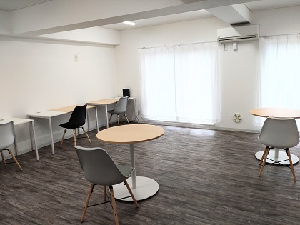 白い壁と木目調グレーのフローリング床の明るい室内に、丸いカフェテーブルと椅子、壁際のカウンターデスクと椅子、白いカーテンの窓が配置されたワーキングスペースの写真