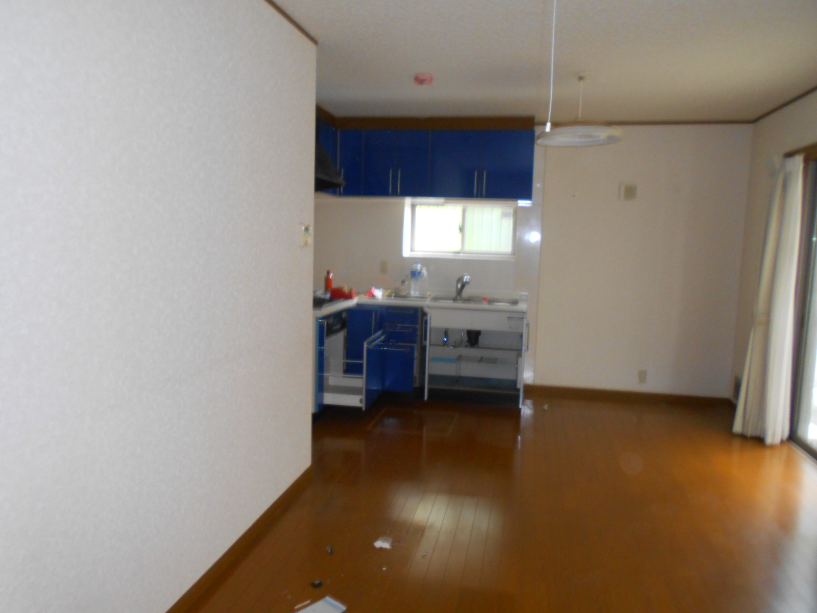 白い壁と木目フローリングの広々としたリビングダイニングキッチンに、青いキャビネットのシステムキッチンと白いシンクが設置され、窓から自然光が差し込む生活感のある居住空間の写真