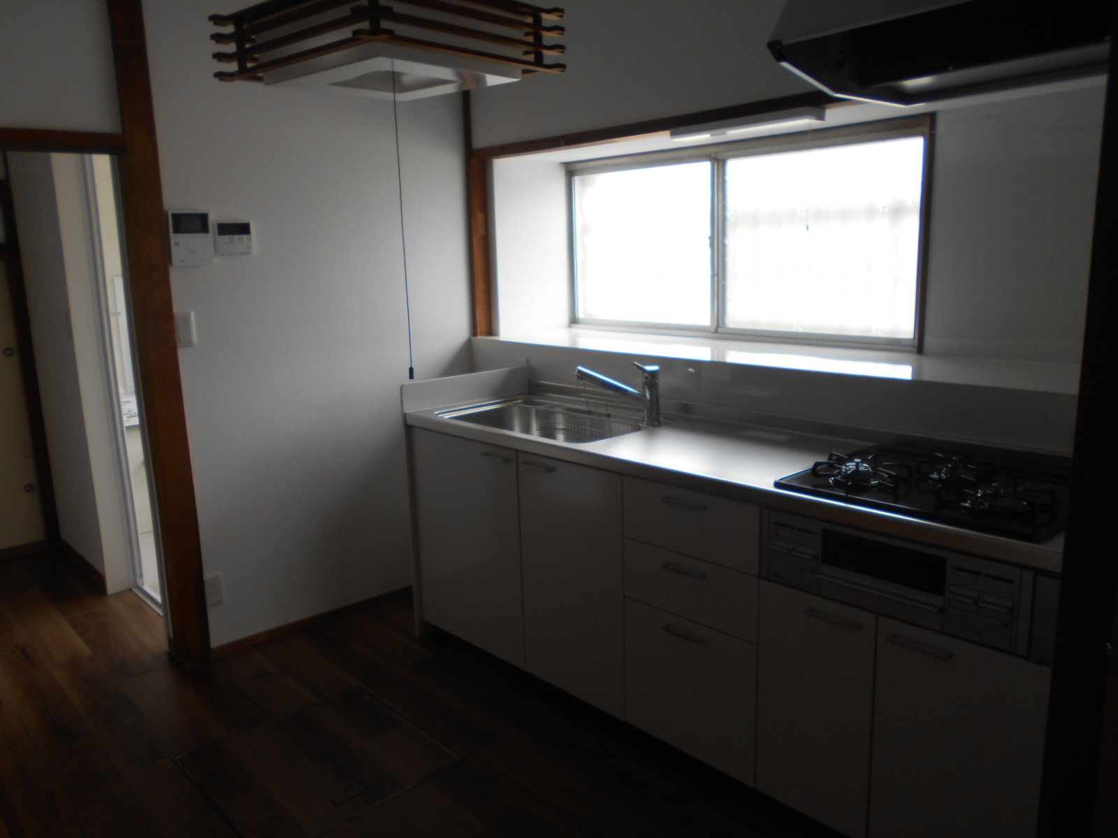 白い壁と木枠の窓が自然光を取り込むL字型キッチンに、ステンレスシンクと黒いガスコンロを備えた白いキャビネットが設置され、和風照明が天井から吊るされたリフォーム済み台所の写真