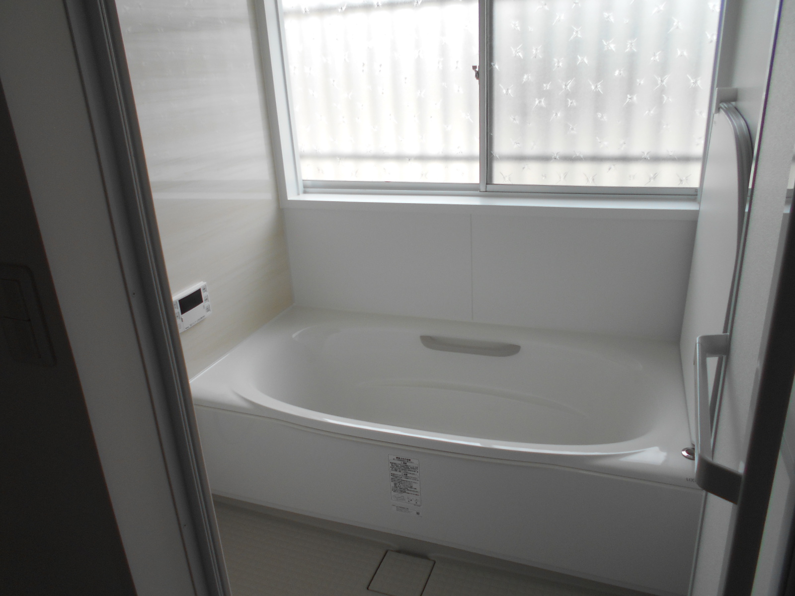 真っ白な壁とユニットバスに擦りガラスの窓から柔らかな光が差し込み、リモコン付き給湯器とシャワーハンドルが設置された清潔感あふれる新築またはリフォーム済み浴室の写真