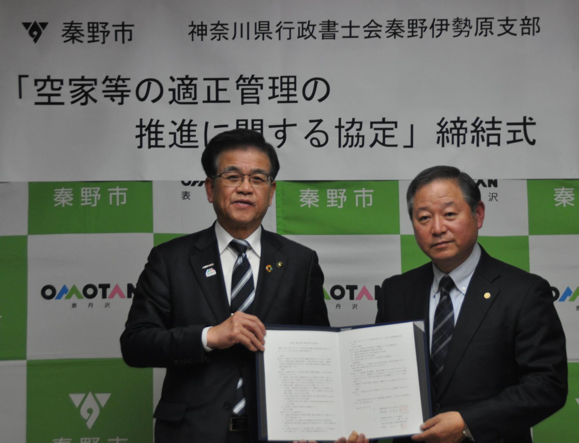 緑と白の秦野市ロゴ入り背景幕の前で「空家等の適正管理の推進に関する協定締結式」と掲げられ、スーツ姿の秦野市長と神奈川県行政書士会秦野伊勢原支部代表が協定書を持ち並ぶ公式調印式の記念写真