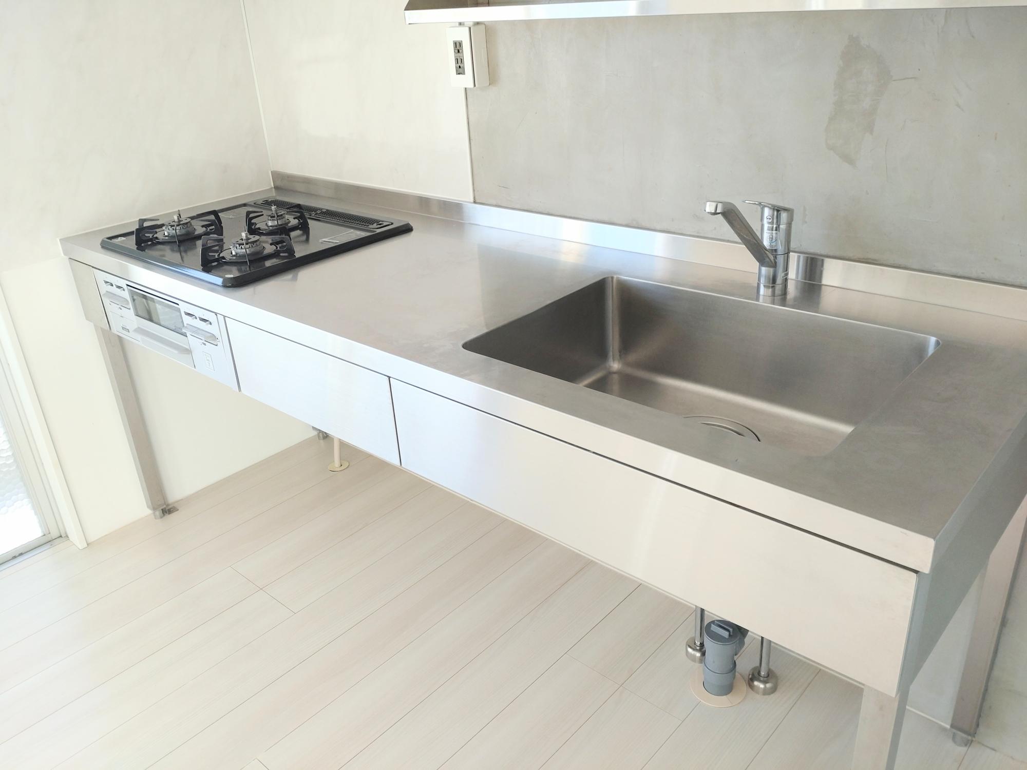 白いタイル床と壁面に、白いキャビネットと人造大理石調のカウンタートップ、左側にガスコンロ、右側に大型ステンレスシンクと水栓が配置された清潔感のあるモダンなシステムキッチンの写真