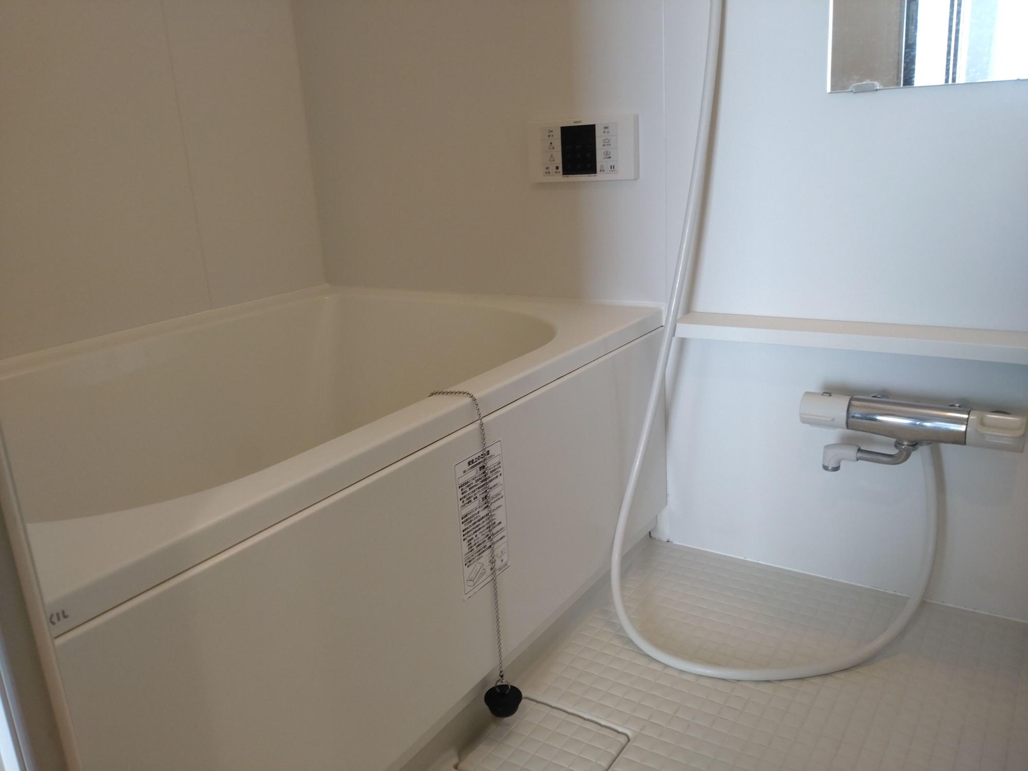 白を基調とした清潔なユニットバスで、左側に浴槽、右側にサーモスタット付きシャワー水栓、壁面にリモコンパネル、右上に小窓が設置された浴室の写真