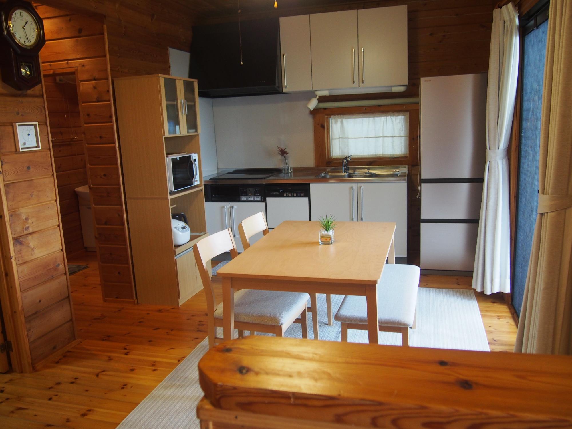 無垢の木材で統一されたログハウス風の室内に白い近代的なキッチンとダイニングテーブルが配置され、振り子時計やカーテンが設置された室内の写真