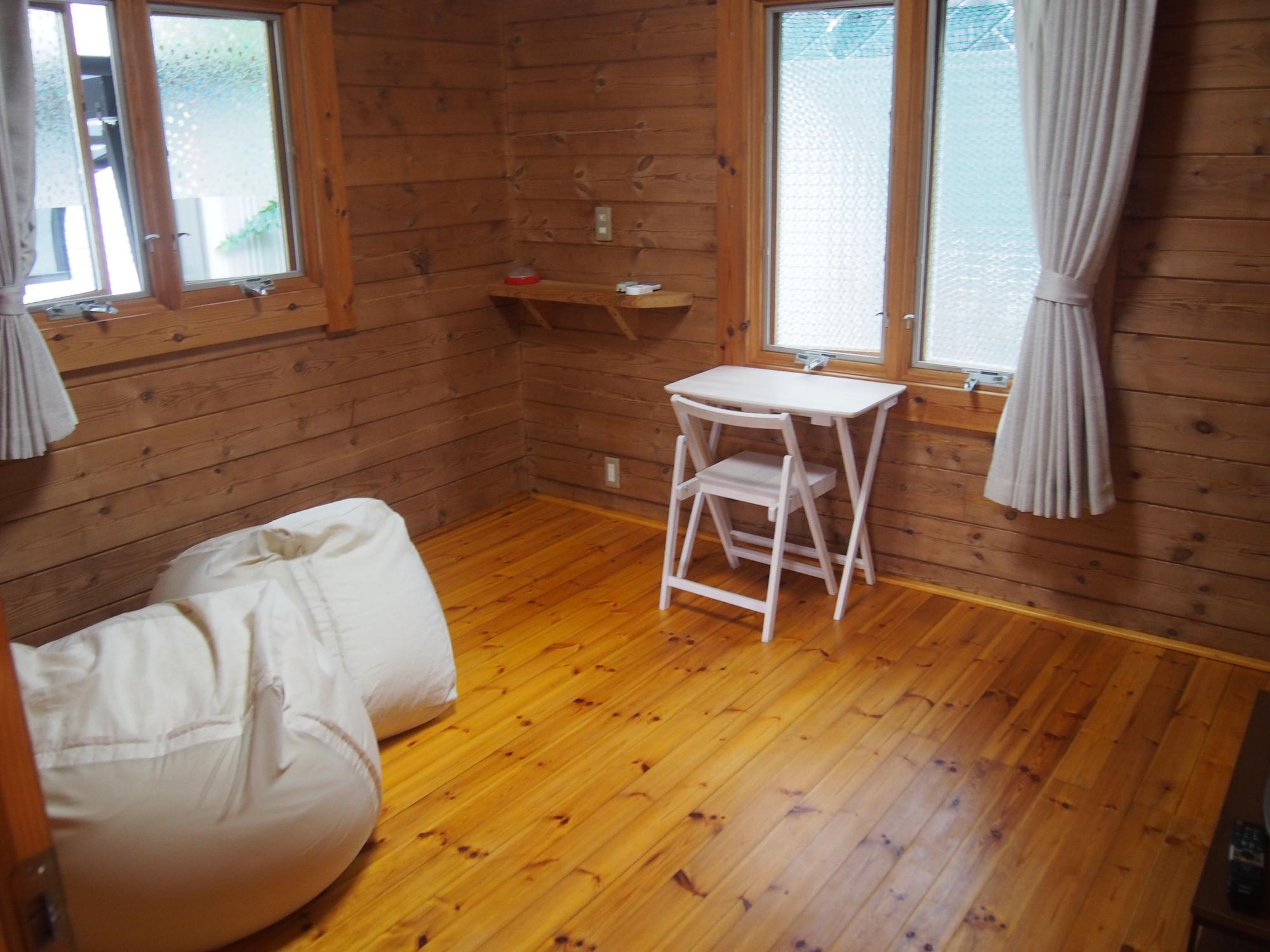 天然木の壁と床で統一された明るい室内に白いビーズクッションと小さな折りたたみ机が配置され、大きな窓から自然光が差し込む室内の写真