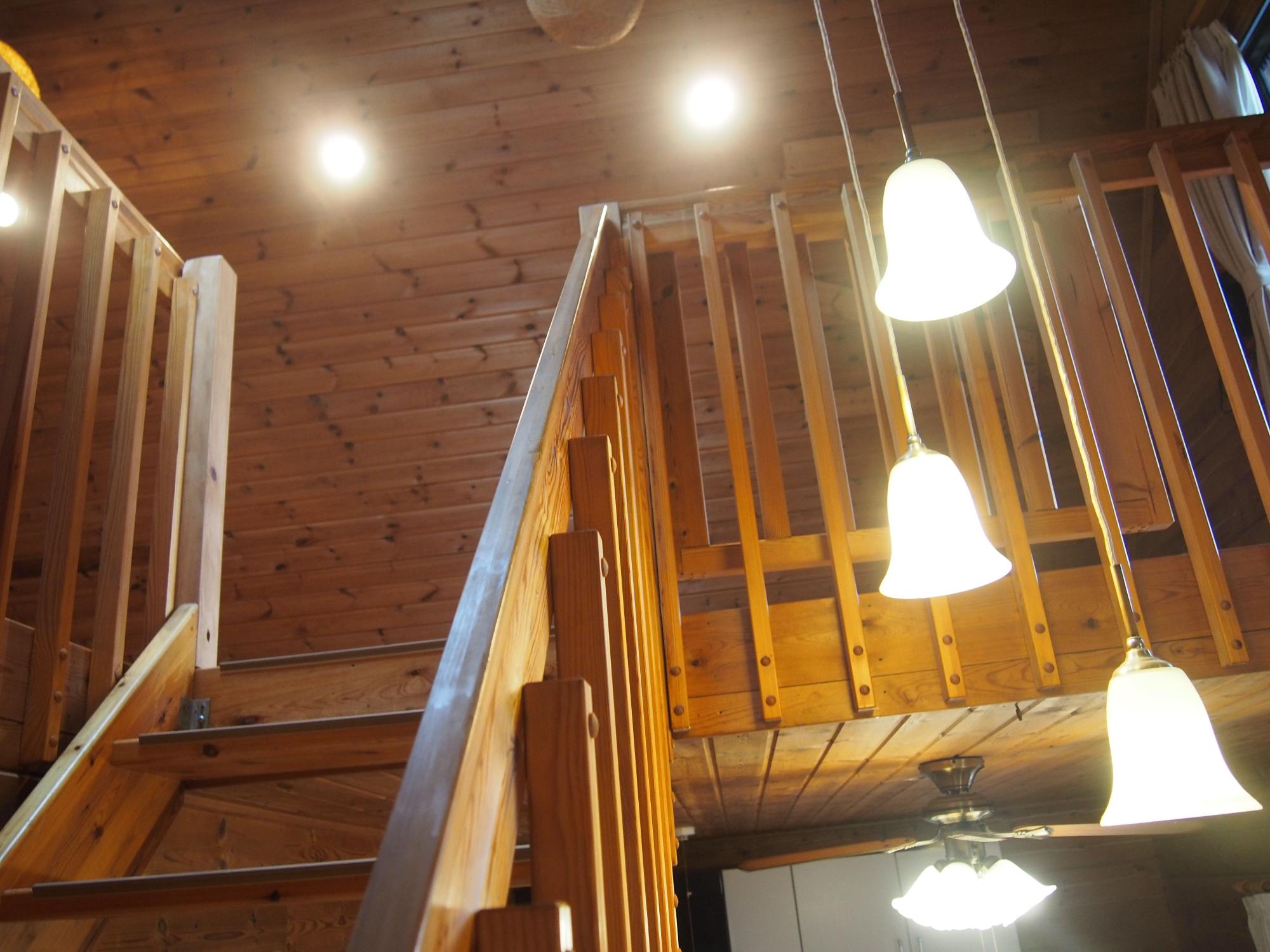 木製の階段と手すりが吹き抜け空間を囲み、複数の白い釣鐘型ペンダントライトが天井から吊り下がるログハウス風住宅の二階部分を見上げた内装写真