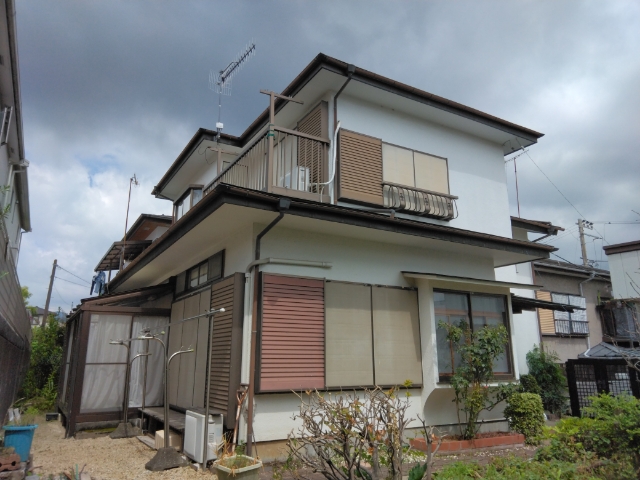 白い外壁に濃い茶色の庇や雨戸、バルコニーの手すりがアクセントとなっている、日本の標準的な二階建て住宅を敷地の庭側から見上げた写真