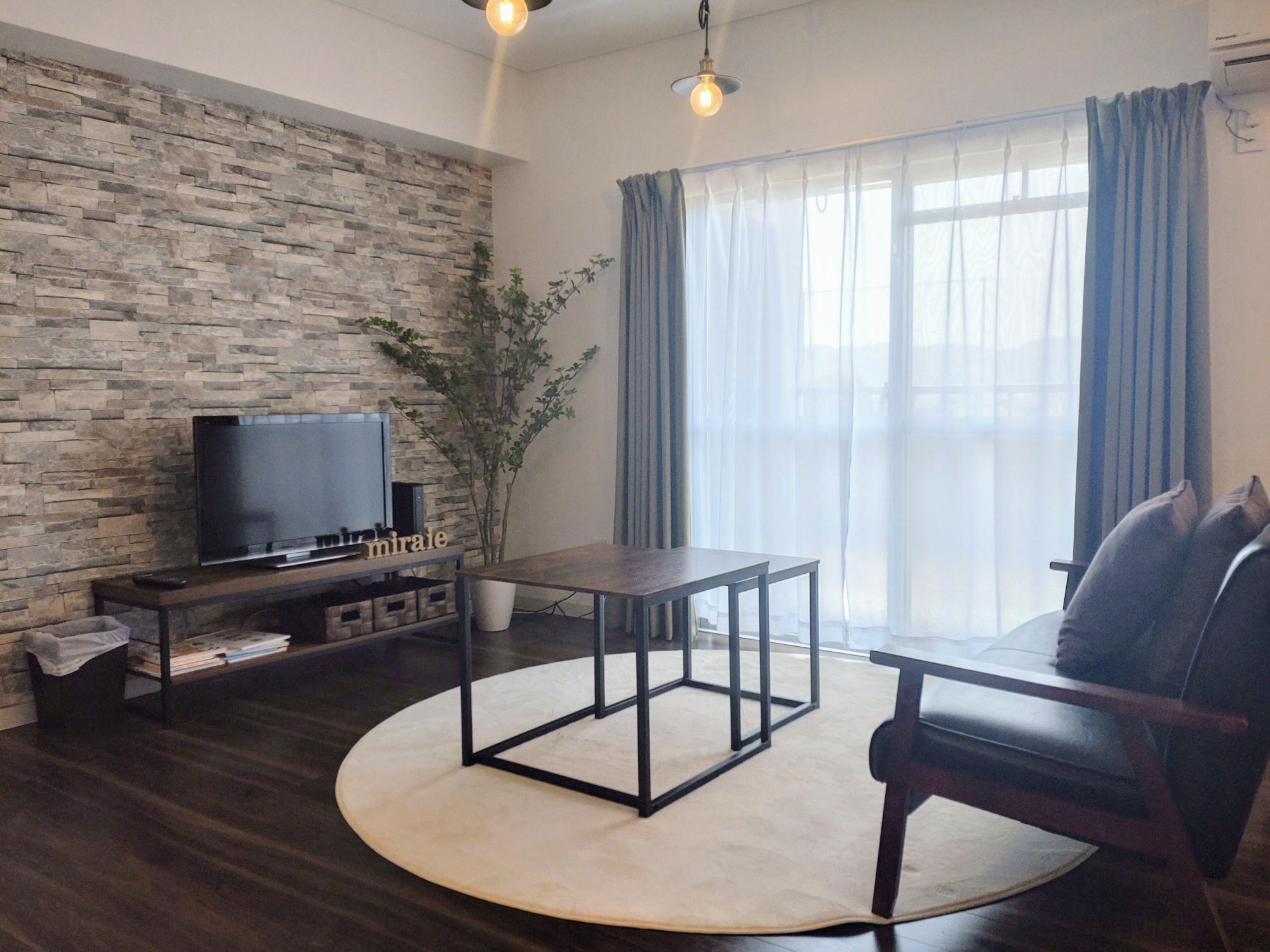 グレーのレンガ調アクセントウォールとテレビボード、黒いフレームのローテーブルが置かれた円形ラグの上に、青いカーテンの窓と茶色いソファが配置されたリビングルームの写真