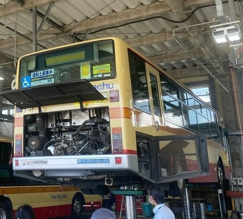 整備工場内でリフトアップされた茶色とオレンジ帯のバスの車体下部メンテナンス作業中の様子を、複数の作業員が点検している様子の写真