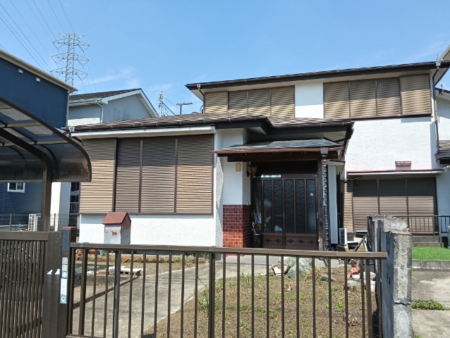 水平ルーバーの茶色い雨戸が特徴的な白壁の二階建て住宅が、フェンスと砂利敷きの空き地越しに見え、送電塔と青空が背景に広がる住宅地の風景写真