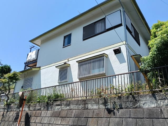 青空を背景に緑に囲まれた高台の白い二階建て住宅が、格子状の手すりとブロック塀の上に立っている写真