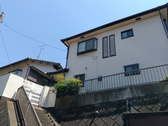 傾斜地に建つ白い二階建て住宅が階段とフェンス越しに見え、左側に給湯器や隣家が並び、電線と青空が広がる住宅密集地の風景写真