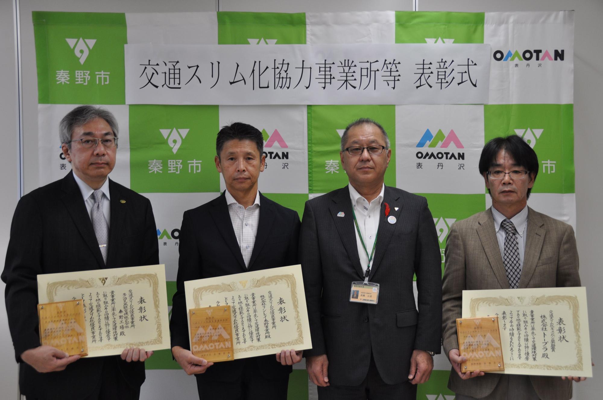 秦野市と表丹沢のロゴが並ぶグリーンと白のバックボードの前で、スーツ姿の4名の男性が表彰状を持って記念撮影している交通スリム化協力事業所等表彰式の写真