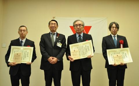 スーツを着た4人の男性が正面を向き、中央の二人が表彰状を持っている、厳かな雰囲気の記念写真