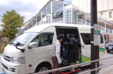 歩道脇に白いワンボックスカーのスライドドアが開いた状態で停車し、利用者が車内へ乗り込む様子を撮影した写真