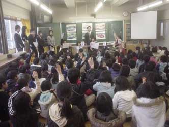 教室の左側に5人の先生、中央に男性の先生が立ち、前に立つ4人の児童や、床に体育座りで座っている多くの児童たちが授業を受け、数人の児童が手を挙げている、特別授業の様子を教室の後方から撮影した写真