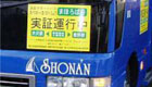 青い車体のバス前面に黄色い文字で「実証運行中」と書かれたステッカーが貼られた企業バスの写真