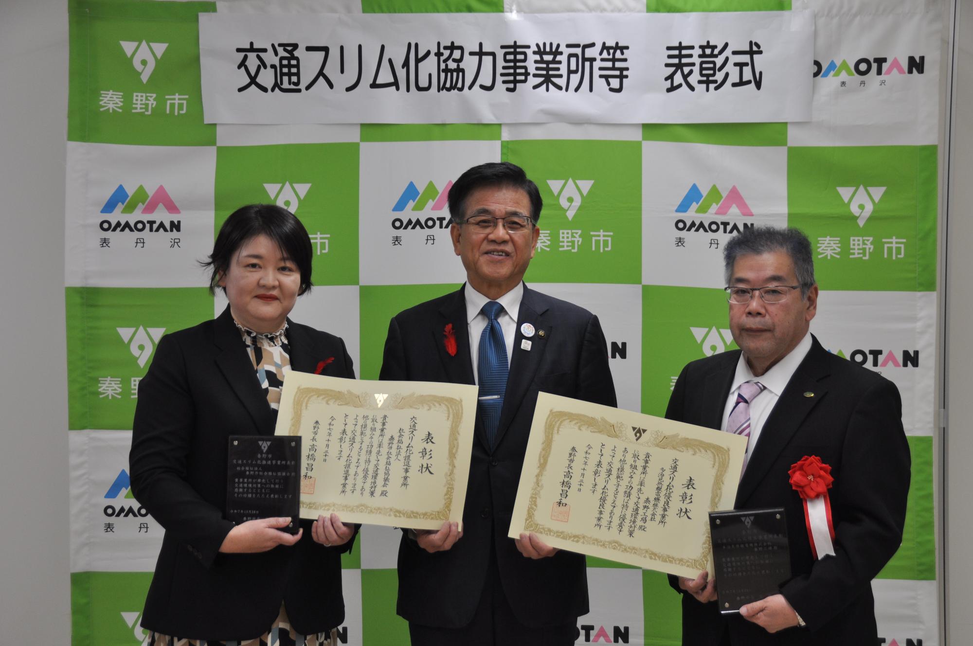 秦野市と表丹沢のロゴが並ぶグリーンと白のバックボードの前で、スーツ姿の4名の男性が表彰状を持って記念撮影している交通スリム化協力事業所等表彰式の写真