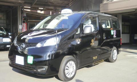 黒塗りの日産NV200バネットと思われるミニバン型のタクシーが、秦野市制施行60周年を記念するロゴを側面に付けて停車している様子の写真