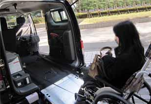 スロープを展開した後部ドアが開いた黒い福祉車両に、黒いコートを着た長い髪の女性が車椅子に乗ったままバッグを膝に置いて乗り込もうとしている様子の写真