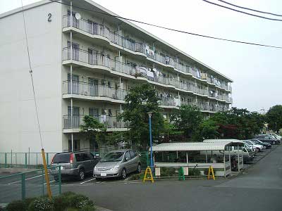 緑色のフェンスで囲まれた敷地に、5階建ての白い集合住宅が建っており、その手前の広いアスファルトの駐車場には多数の車が駐車され、一部に屋根付きの駐輪場もある市営薬師原団地の写真